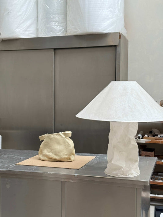 Handcrafted ceramic vase object with unique texture and form displayed on a metal table next to a paper-textured lamp in an artisan studio setting