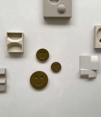 Olive 'HAPPY' ceramic artwork by Edith Beurskens displayed on wall.