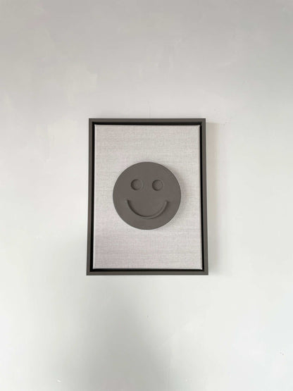 Canvas artwork with a smiley face design, encased in a black frame, made from biodegradable PLA and finished with plaster.