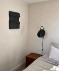 3D art piece '#22.03 Cécé' on a wall above a wooden nightstand in a minimalist bedroom setting.