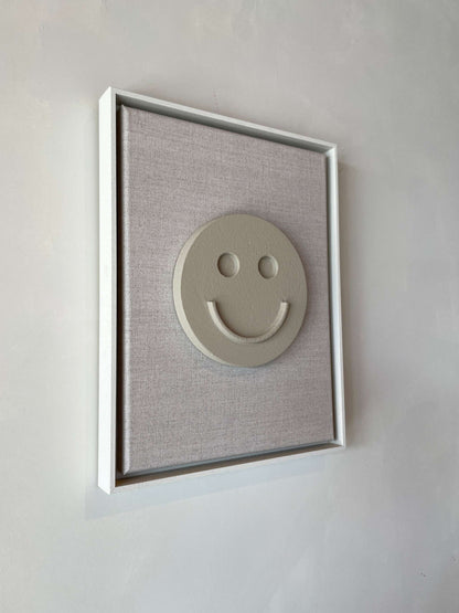 Canvas Happy artwork with a smiley face on brown wood, framed in shadow white, made from biodegradable PLA and finished with structured plaster.