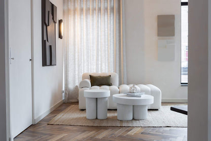Modern living room featuring a white sculptural armchair, two round white side tables, abstract wall art, and textured curtains.
